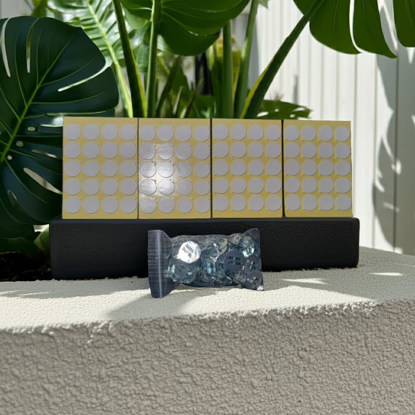 A bag of 60 wick holders and 4 sheets of 80 heat-proof stickers on a textured surface with plants in the background