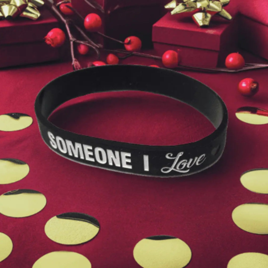 Black 'SOMEONE I Love' wristband on a red surface with gold polka dots and red berries.