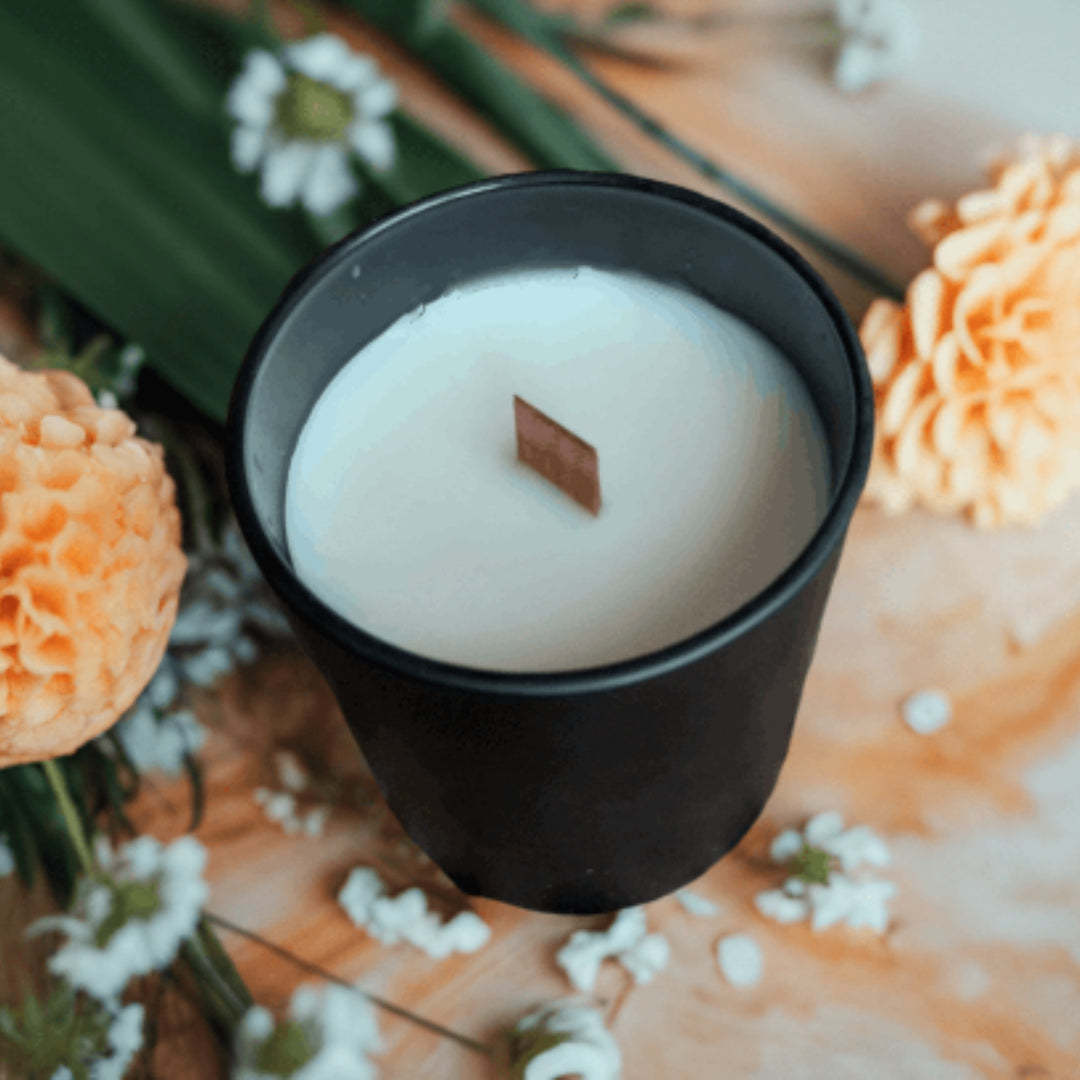 White candle in a black container surrounded by flowers on a wooden surface