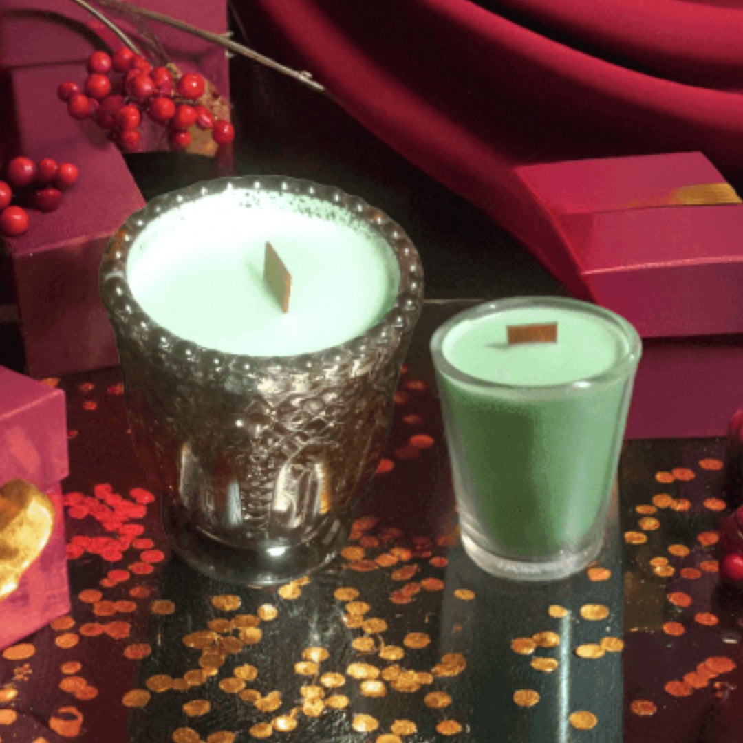 Two small candles in glass holders on a decorative surface with red berries and gift boxes.