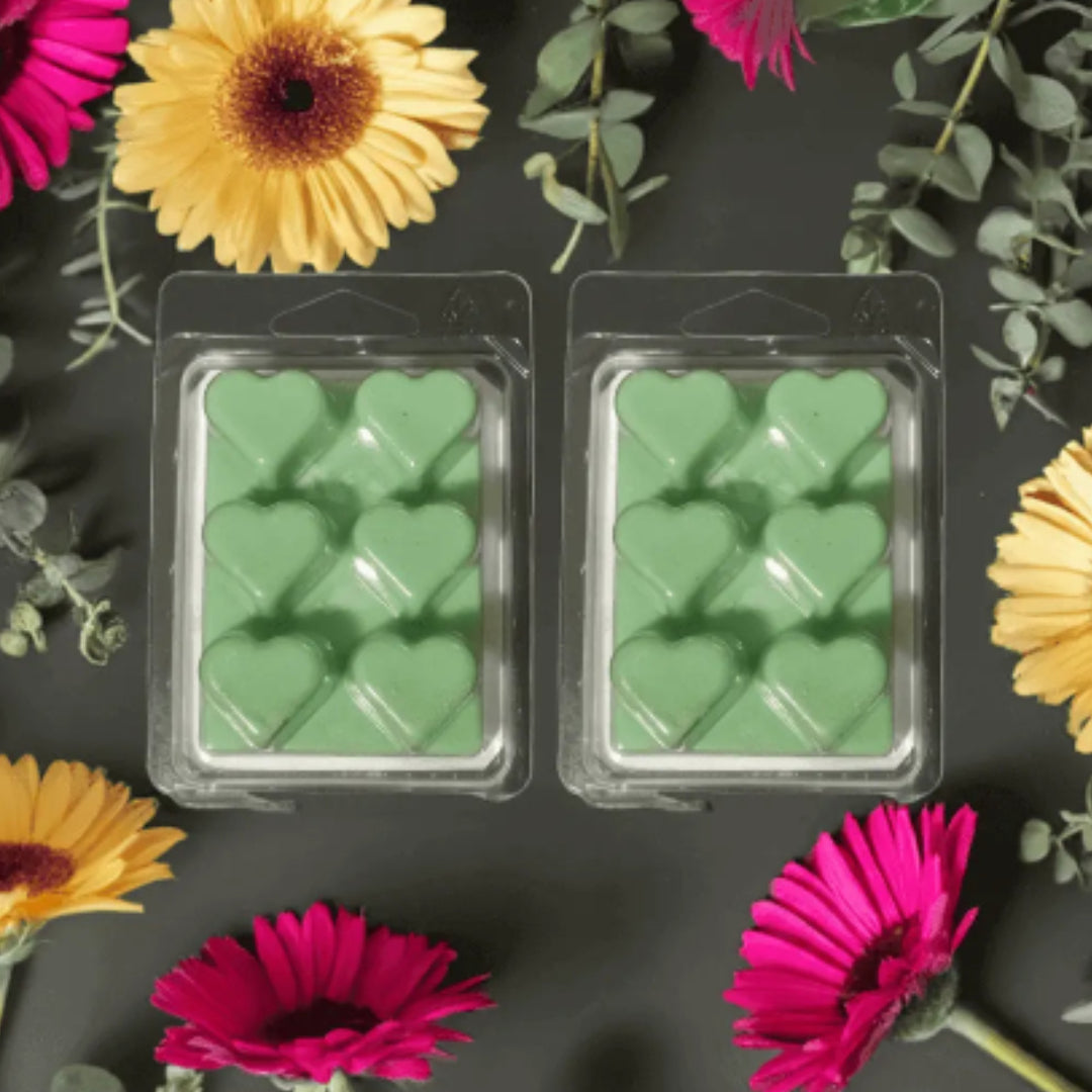 Two packages of green heart-shaped wax melts surrounded by colorful flowers on a dark background