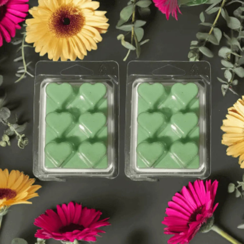 Two packages of green heart-shaped wax melts surrounded by colorful flowers on a dark background