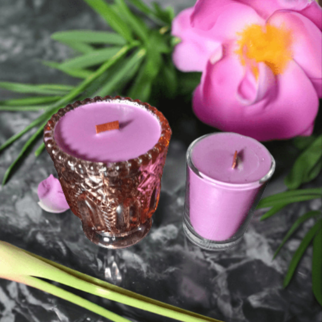 Two purple candles in glass holders on a dark surface with green leaves and a pink flower.