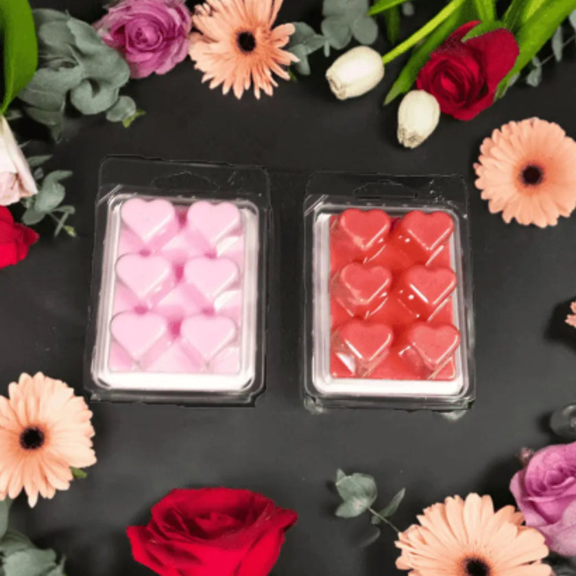 Two packages of heart-shaped wax melts surrounded by flowers on a dark surface