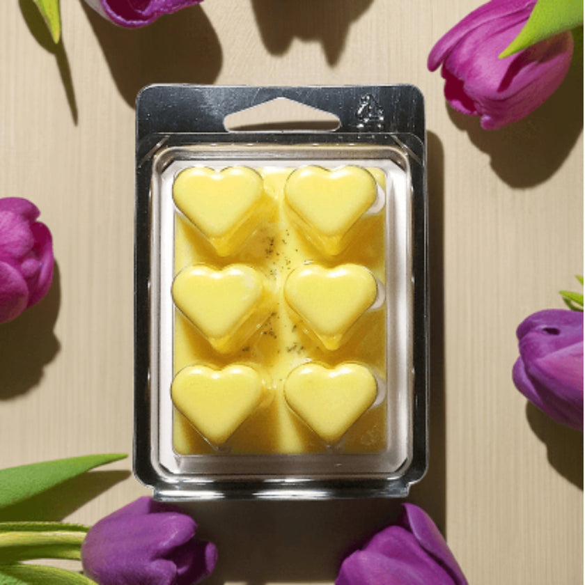 Packaged yellow heart-shaped wax melts on a beige surface with purple tulips.