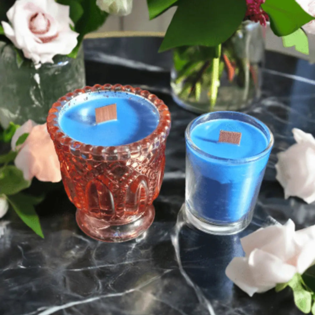 Two glass candles, one red and one blue, on a dark surface with flowers in the background.