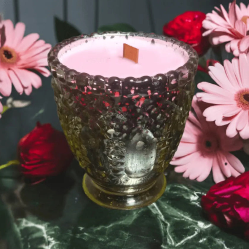 Pink candle in a decorative glass holder surrounded by pink and red flowers on a green surface