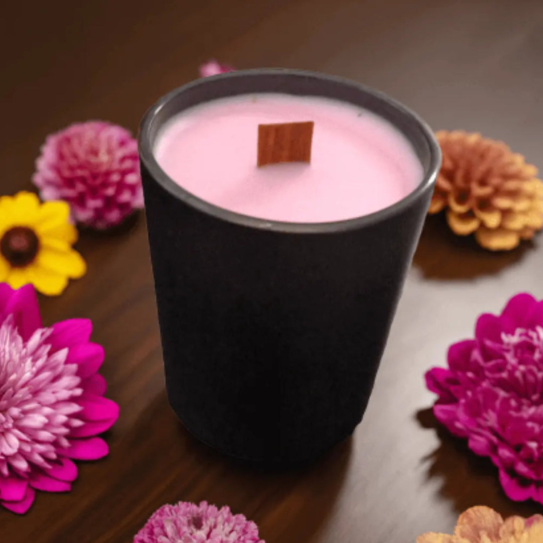 Pink candle in a black container with a wooden wick, surrounded by colorful flowers on a wooden surface.