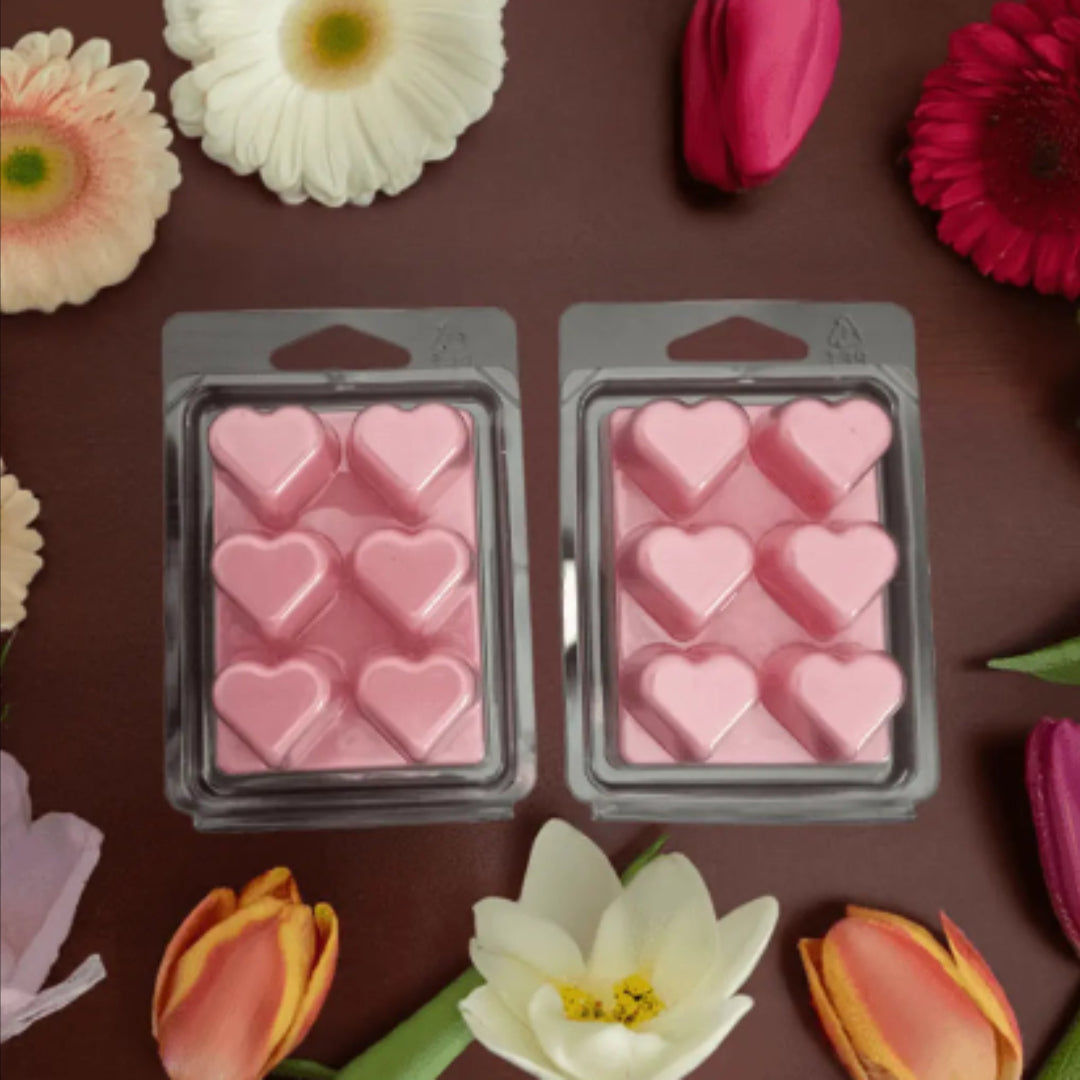 Two packages of pink heart-shaped wax melts on a brown surface with flowers around.